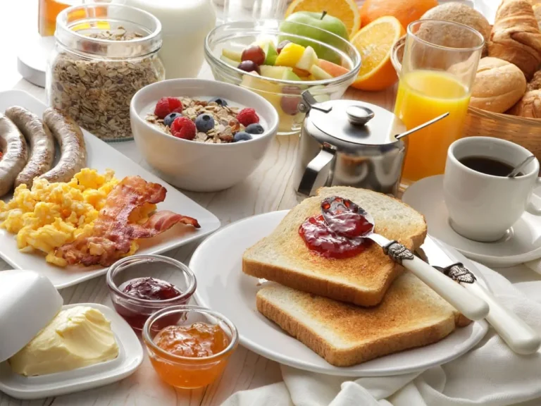 A lavish breakfast spread featuring scrambled eggs, bacon, sausage, toast with jam, yogurt with berries, granola, fruit salad, orange juice, and coffee.